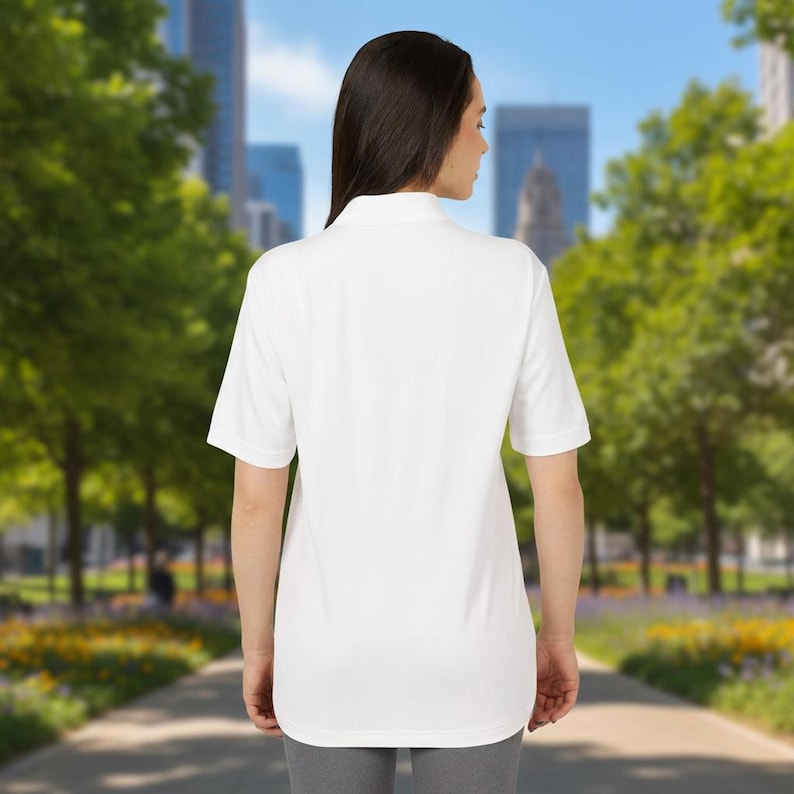 A woman stands outdoors with her back to the camera, wearing a plain white short-sleeve polo shirt and grey pants.