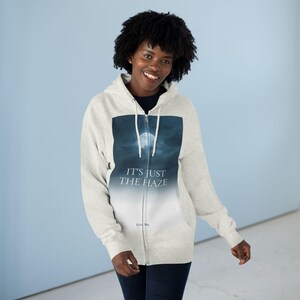 A light grey zip-up hoodie with a graphic of a full moon in a dark blue sky and the text "IT'S JUST THE HAZE". The hoodie has a drawstring hood and a full-length zip.