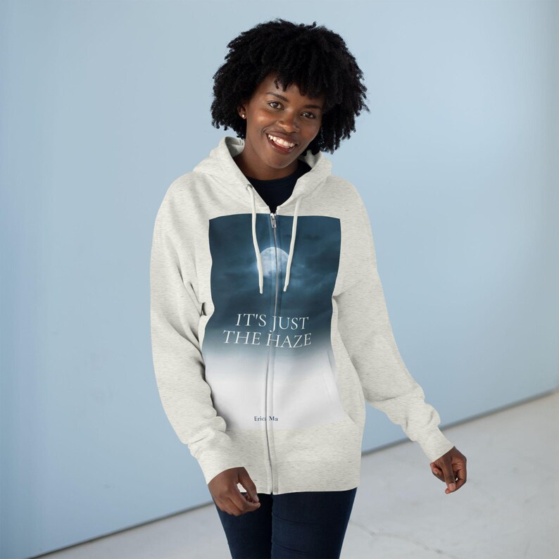 A light grey zip-up hoodie with a graphic of a full moon in a dark blue sky and the text "IT'S JUST THE HAZE". The hoodie has a drawstring hood and a full-length zip.