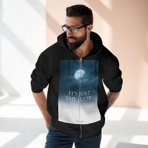 Black zip-up hoodie with a full moon graphic and the text "IT'S JUST THE HAZE". The graphic is set against a dark blue and white gradient background. The hoodie has a drawstring hood and a full-length zip.