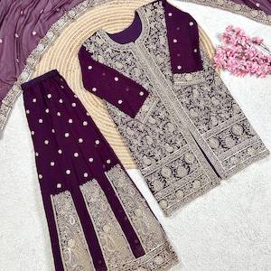 May include: A formal, plum-colored ensemble featuring a long-sleeved jacket and matching pants, both adorned with intricate gold embroidery. A sheer scarf with delicate embellishments completes the outfit. The set is displayed on a neutral surface.