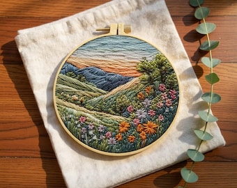 May include: A detailed embroidered landscape scene within a wooden hoop. The artwork showcases green hills, a variety of wildflowers, and a sunset with orange and blue tones. The embroidery is set in a wooden hoop and rests on a cream-coloured fabric.