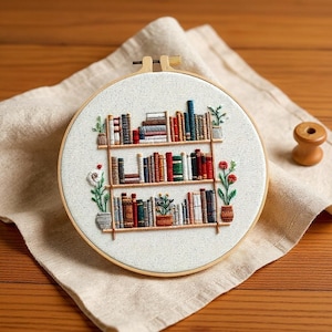 以下が含まれることがあります： 円形に刺繍されたアート作品。3段の棚がある本棚が描かれています。棚には様々な色の本が並び、小さな鉢植えの植物も配置されています。背景は質感のあるオフホワイトの生地で、すべて木製のフレームに収められています。