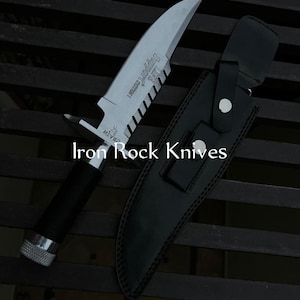 Puede incluir: Un cuchillo de acero inoxidable con filo dentado y mango negro, acompañado de una funda de cuero negro. El cuchillo tiene el texto "LIFE Support System 1" y "Iron Rock Knives" en la hoja.