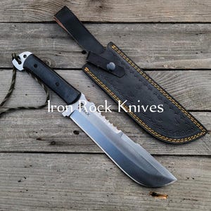 May include: A large, silver-bladed knife with a black handle and a black leather sheath. The knife has a serrated edge near the handle and a lanyard. The sheath is embossed with a pattern and has yellow stitching.
