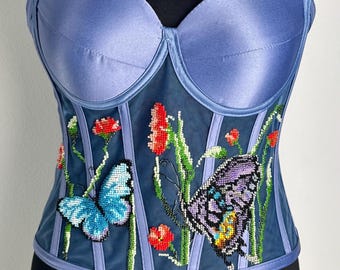 May include: A periwinkle-colored corset with a sweetheart neckline. The bodice features beaded butterflies and floral embroidery. Vertical stripes and a satin finish add detail to this strapless top.