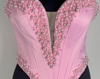 May include: A light pink corset with a deep V-neckline, embellished with rows of small, pearlescent beads. The corset is displayed on a black mannequin, showcasing its structured design and satin fabric.