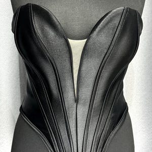 May include: A black strapless corset with a structured design. The corset features vertical paneling and a deep V-neckline. The garment is displayed on a black mannequin, showcasing its form-fitting silhouette and satin-like texture.