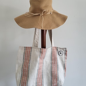 May include: A wide-brimmed straw sun hat and a beige tote bag with red and grey stripes. The tote bag features a small black patch with a white "K" embroidered on it. The hat has a string tied around the crown.