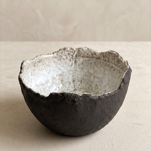 Pode incluir: Uma pequena tigela redonda com um exterior preto mate escuro e um interior branco contrastante salpicado com pontos castanhos. A borda da tigela apresenta um contorno irregular, serrilhado e texturizado, conferindo-lhe um aspeto bruto.