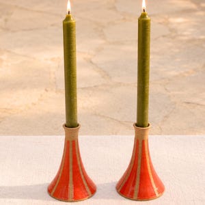 May include: Two ceramic candle holders with a flared base and a vibrant orange glaze featuring vertical unglazed tan stripes. Each holder supports a tall, slender olive green taper candle with a lit flame, creating a warm and balanced display.