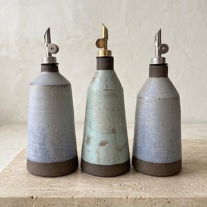 Puede incluir: Tres botellas de aceite de cerámica con boquillas de acero inoxidable y latón. Las botellas tienen una combinación de colores azul claro, verde y gris con una base marrón oscuro. Las botellas están dispuestas en fila.