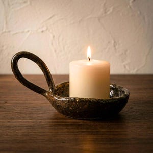 Puede incluir: Una vela encendida de color marfil en un candelero de cerámica marrón oscuro con asa. El candelero tiene una superficie texturizada y moteada, colocado sobre una superficie de madera. La llama es brillante.