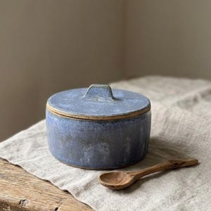 Puede incluir: Un tarro de cerámica azul con tapa y una pequeña cuchara de madera. El tarro es redondo con un asa en la tapa. La cuchara es pequeña y descansa sobre un paño de lino. El tarro y la cuchara están sobre una mesa de madera.