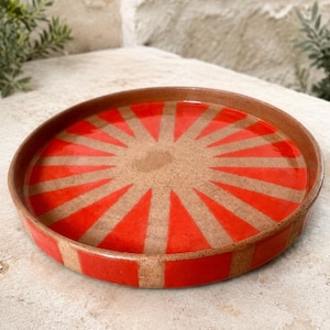Puede incluir: Una bandeja redonda de cerámica con una base de color tostado moteado y un vibrante patrón de rayos rojos que irradia desde el centro. El diseño consta de segmentos triangulares alternos en rojo y tostado, con el patrón rojo continuando por el borde.