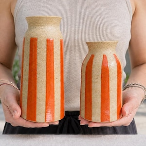 Op de afbeelding: Twee keramische vazen met een gespikkelde beige afwerking en verticale oranje strepen. De vazen hebben een cilindrische vorm met een licht uitlopende rand. De langere vaas staat links en de kortere vaas rechts, beide met een gladde textuur.