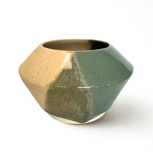 May include: A two-toned ceramic bowl with a geometric design. The bowl is divided diagonally, with one side in a speckled tan and the other in a glossy green. The interior is a darker shade of brown. The bowl sits on a white base.
