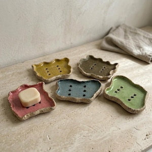 Puede incluir: Una colección de cinco jaboneras de cerámica en varios colores: rosa, amarillo, verde, azul y marrón. Cada plato tiene una forma orgánica única con un borde moteado y texturizado y orificios de drenaje. Un plato rosa contiene una pastilla de jabón.