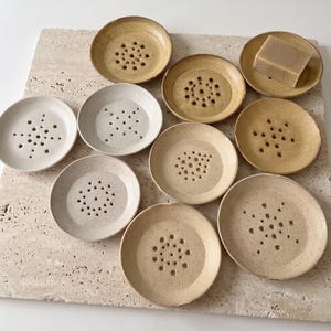 May include: A collection of nine round ceramic soap dishes in various shades of beige, tan, and off-white, each featuring a central pattern of drainage holes. One dish holds a rectangular bar of light brown soap, all arranged on a textured surface.