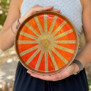 May include: A round ceramic tray featuring a vibrant sunburst pattern with alternating rays of bright orange and muted gold. The tray has a raised rim and a smooth, glossy finish, showcasing a radial design that extends from the center outward.