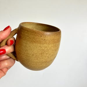 Könnte beinhalten: Handgefertigte Keramiktasse mit gesprenkelter Oberfläche in einem warmen, erdigen Farbton. Die Tasse hat eine abgerundete Form und einen stabilen Henkel. Innen und außen haben eine ähnliche Farbe, mit Variationen in der Glasur. Ideal für Kaffee oder Tee.