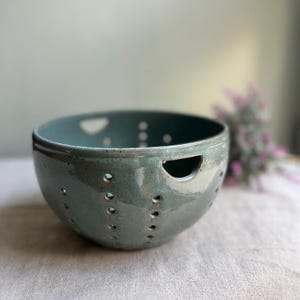 May include: A teal ceramic bowl with a series of small, evenly spaced holes around the sides. The bowl has a rounded shape with a slightly flared rim and a cutout on one side. The background is blurred, with a hint of purple flowers.