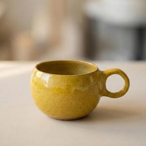 Könnte beinhalten: Eine kleine, runde, gelbe Keramik-Tasse mit Henkel. Die Tasse hat eine gesprenkelte Textur und eine glänzende Oberfläche. Der Innenraum ist etwas dunkler gelb. Die Tasse ist für Getränke konzipiert.