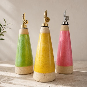 Puede incluir: Tres dispensadores de aceite cónicos de cerámica con bases beige moteadas y secciones centrales esmaltadas en colores vibrantes verde, amarillo y rosa. Cada botella cuenta con un vertedor metálico con tapa, dos en oro y uno en plata.