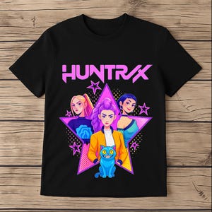 May include: Black t-shirt with a graphic design. The design features the word "HUNTRAX" in pink, three stylized female figures, a blue cat, and a pink star. The design has a cartoonish, futuristic aesthetic.