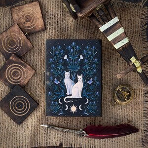Puede incluir: Un diario azul oscuro con un dise&ntilde;o celestial con dos gatos blancos, flores azules, mariposas y lunas crecientes. El diario est&aacute; rodeado de posavasos de madera, una br&uacute;jula y una pluma con una pluma roja.