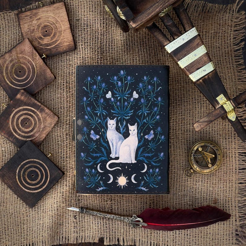 Puede incluir: Un diario azul oscuro con un dise&ntilde;o celestial con dos gatos blancos, flores azules, mariposas y lunas crecientes. El diario est&aacute; rodeado de posavasos de madera, una br&uacute;jula y una pluma con una pluma roja.