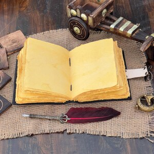 Puede incluir: Un diario abierto de estilo antiguo con p&aacute;ginas amarillentas y envejecidas descansa sobre una superficie de arpillera. Tambi&eacute;n hay un bol&iacute;grafo decorativo con una pluma roja y un peque&ntilde;o carro de madera. El diario est&aacute; encuadernado en cuero negro.