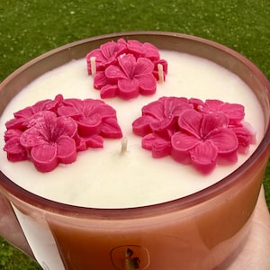 Puede incluir: Una vela en un tarro de cristal color ámbar relleno de cera blanca. La parte superior presenta tres grupos de decoraciones de cera moldeada en forma de hibisco rosa brillante y tres mechas. Una etiqueta dorada muestra el texto MKCANDLE.