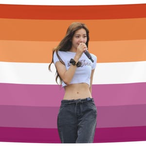 Puede incluir: Una persona sostiene un micrófono frente a una bandera del orgullo lésbico. La bandera tiene franjas horizontales de color naranja, blanco, rosa y morado. La persona lleva un top corto blanco y vaqueros oscuros.