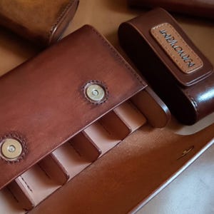 Puede incluir: Primer plano de varias fundas de armónica de cuero marrón. Una funda está abierta, revelando ranuras interiores. Otra funda está cerrada, con la palabra "HARMONICA" grabada en un pequeño parche rectangular. Las fundas son de cuero marrón oscuro.