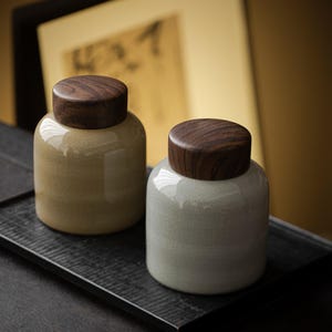 Può includere: Due barattoli in ceramica con coperchi in legno. Uno è beige, l'altro grigio chiaro. Entrambi sono cilindrici con la parte superiore arrotondata, su un vassoio nero. I coperchi in legno marrone scuro sono circolari.
