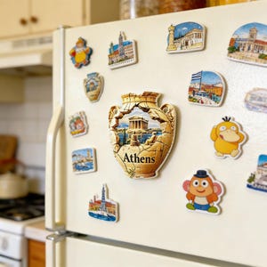 Puede incluir: Una colección de coloridos imanes para nevera, incluyendo un imán grande en forma de jarrón agrietado con "Atenas" impreso. Otros imanes representan monumentos y personajes de dibujos animados. Los imanes están adheridos a la puerta blanca de una nevera.