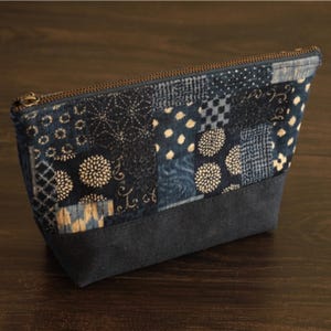 Puede incluir: Un neceser patchwork azul marino con cremallera marrón oscuro. La bolsa presenta una variedad de cuadrados de tela estampados en tonos azules y blancos, con una base azul marino liso.