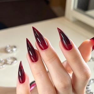 Op de afbeelding: Lange, stiletto-vormige vingernagels met een diep, doorschijnend rood kleurverloop dat overgaat in een donkerdere, bijna zwarte tint bij de punten. De nagels hebben een glanzende, gladde afwerking die het licht reflecteert voor een scherpe look.