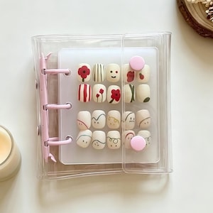 May include: A clear, pink-hinged case holding a set of press-on nails. The nails feature various designs, including floral patterns, stripes, and abstract art. A lit candle and a wooden coaster with dried flowers are in the background.