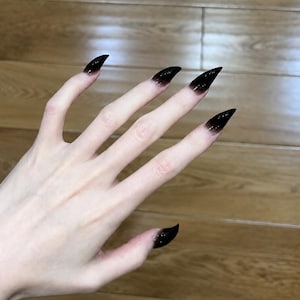May include: A hand with long, pointed, stiletto-shaped artificial nails. The nails feature a gradient design, transitioning from a clear, translucent base near the cuticle to a solid, opaque black color at the sharp, tapered tips of each nail.