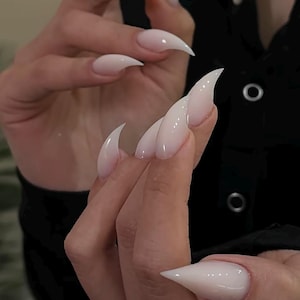 Puede incluir: Un conjunto de uñas postizas largas en forma de estilete con un acabado degradado de blanco lechoso a rosa pálido. Las uñas presentan una punta afilada y una superficie brillante, mostrando un aspecto limpio y uniforme en todos los dedos visibles.