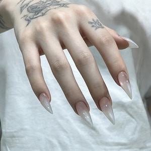 Puede incluir: U&ntilde;as artificiales en forma de estilete con un degradado ombr&eacute; blanco lechoso transl&uacute;cido que se desvanece hacia un acabado transparente y brillante en las puntas. Las u&ntilde;as tienen una silueta afilada y puntiaguda y una superficie lisa y reflectante.
