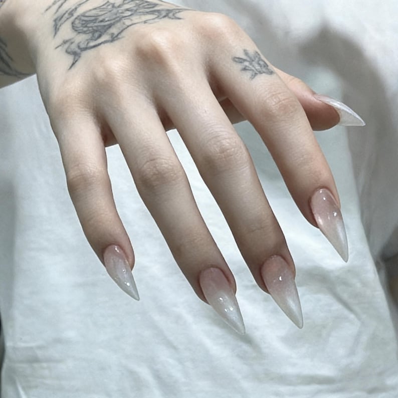 Puede incluir: U&ntilde;as artificiales en forma de estilete con un degradado ombr&eacute; blanco lechoso transl&uacute;cido que se desvanece hacia un acabado transparente y brillante en las puntas. Las u&ntilde;as tienen una silueta afilada y puntiaguda y una superficie lisa y reflectante.