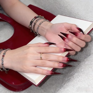 Puede incluir: Manos con uñas largas y puntiagudas, estilo ombre, que van del transparente al rojo intenso. Un anillo de oro adorna un dedo. Las manos descansan sobre un libro abierto y están adornadas con múltiples pulseras de plata. Un bolso rojo está en el fondo.