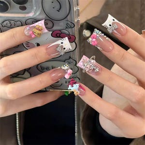 May include: Long, decorated fingernails with various designs. Some nails feature Hello Kitty characters, bows, and rhinestones. Others have a clear base with embellishments, including a small unicorn and other decorative elements.