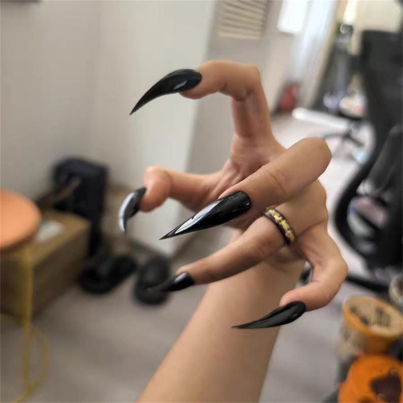 May include: A hand with long, pointed, black acrylic nails. The nails are glossy and sharp, creating a dramatic effect. A gold ring with black accents is on the ring finger.