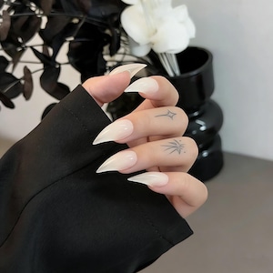 Puede incluir: Uñas acrílicas largas, puntiagudas y translúcidas. Las uñas son de un color pálido y natural, con una forma afilada de estilete. La mano también tiene dos pequeños y delicados tatuajes en los dedos. Las uñas son un accesorio de moda.
