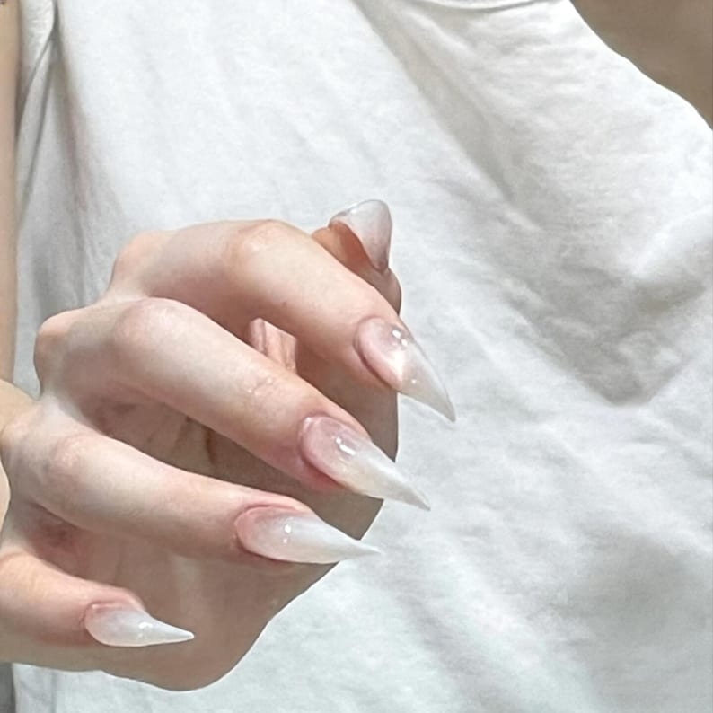 Puede incluir: U&ntilde;as postizas largas en forma de estilete puntiagudo con un acabado degradado blanco lechoso y transl&uacute;cido. Las u&ntilde;as presentan una superficie lisa y brillante que transiciona desde una base transparente hacia una punta blanca opaca.