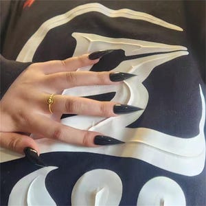 May include: A hand with long, pointed black fingernails. A gold ring with a small, round design is on the ring finger. The hand rests on a black garment with a white graphic design.